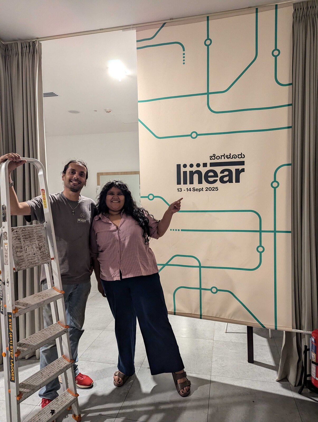 Two of the team beside a tall metro-line banner for Linear Bengaluru, 13 to 14 September 2025, during the festival install