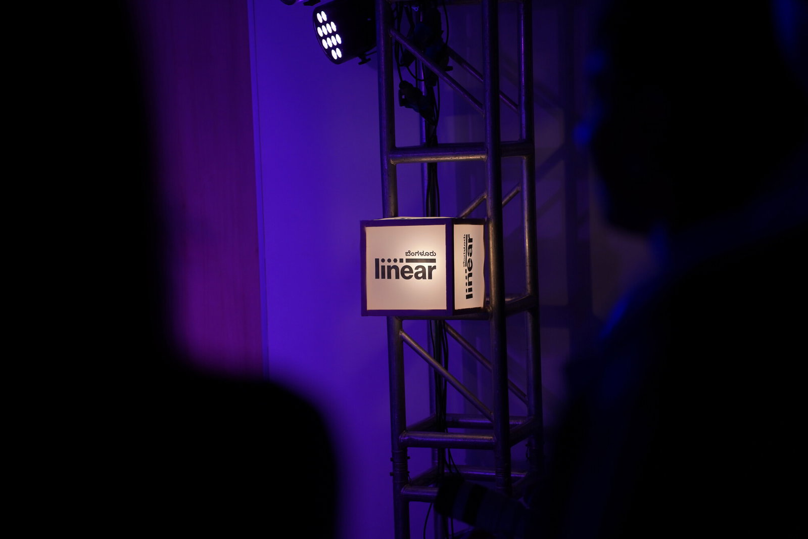 A Linear Bengaluru lightbox cube glowing on a lighting truss at an event, the wordmark on one face and the Kannada brandline on the other
